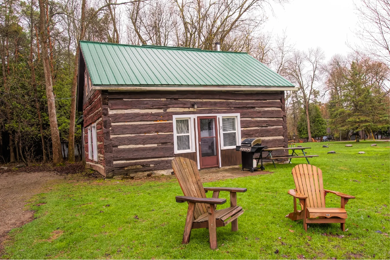 Pigeon Lake Resort Cottage Seven.&nbsp; Exterior of large four-bedroom century log cabin with green metal roof, lawn chairs, barbecue, and picnic table.